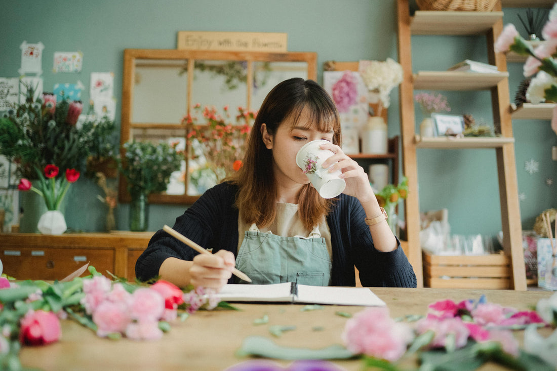 Young woman sitting at a wooden table with flowers and a notebook drinking tea out of a cup