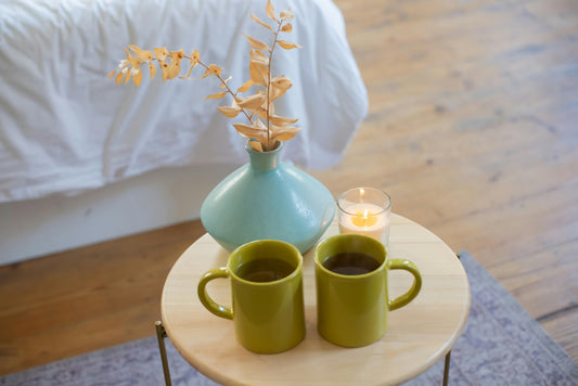 Two green mugs of matcha tea on a side table with a blue potted flower and a candle.