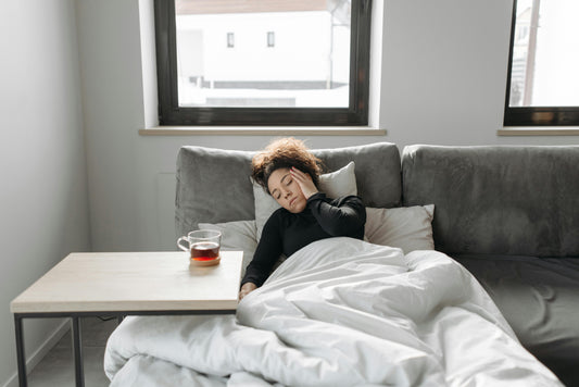Woman lying down on comfy sofa with pillows and a blanket, experiencing cough cold flu symptoms with a warm tea for soothing comfort and symptoms