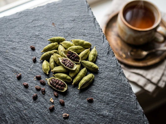Whole green cardamom pods split open to reveal dark seeds on a slate surface with a warm mug of chai blurred in the background