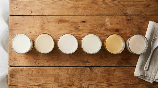 Six small glasses of different milk alternatives arranged in a row on a wooden surface in soft morning light