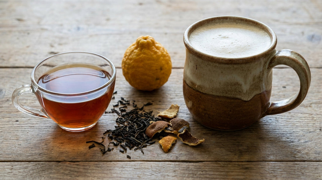A glass teacup of plain Earl Grey tea beside a creamy London Fog latte in a ceramic mug with loose tea leaves and a bergamot fruit between them