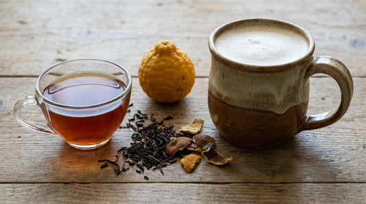 A glass teacup of plain Earl Grey tea beside a creamy London Fog latte in a ceramic mug with loose tea leaves and a bergamot fruit between them