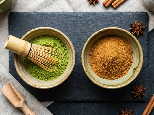 Two small ceramic bowls side by side on dark slate, one with bright green matcha powder and one with warm amber chai powder, bamboo whisk resting across the matcha bowl