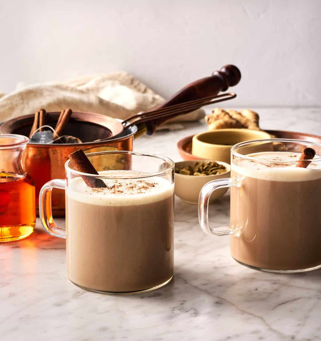 chai lattes in glass cups with cinnamon sticks in them and powdered brown sugar on top, with ingredients and utensils in the background