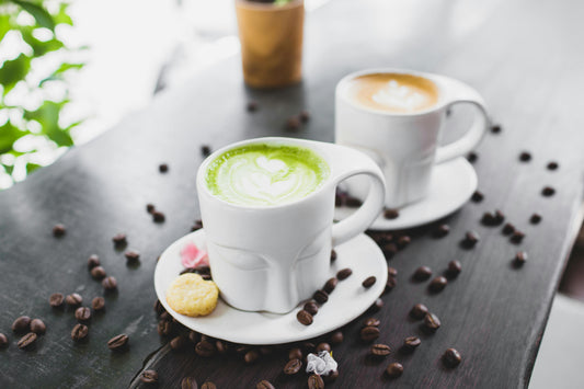 A mug of matcha latte beside a mug of coffee latte on a wooden table with coffee beans
